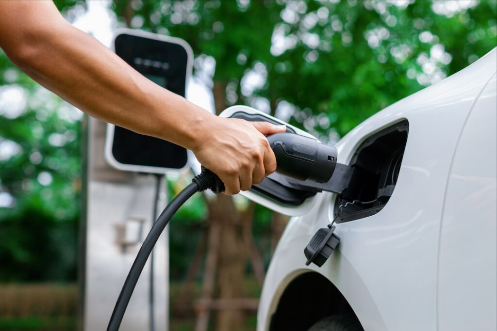 Hand inserting EV charging plug to electric vehicle in focus shot with blurred background of outdoor natural greenery. Progressive sustainable energy powered electric charging station for rechargeable